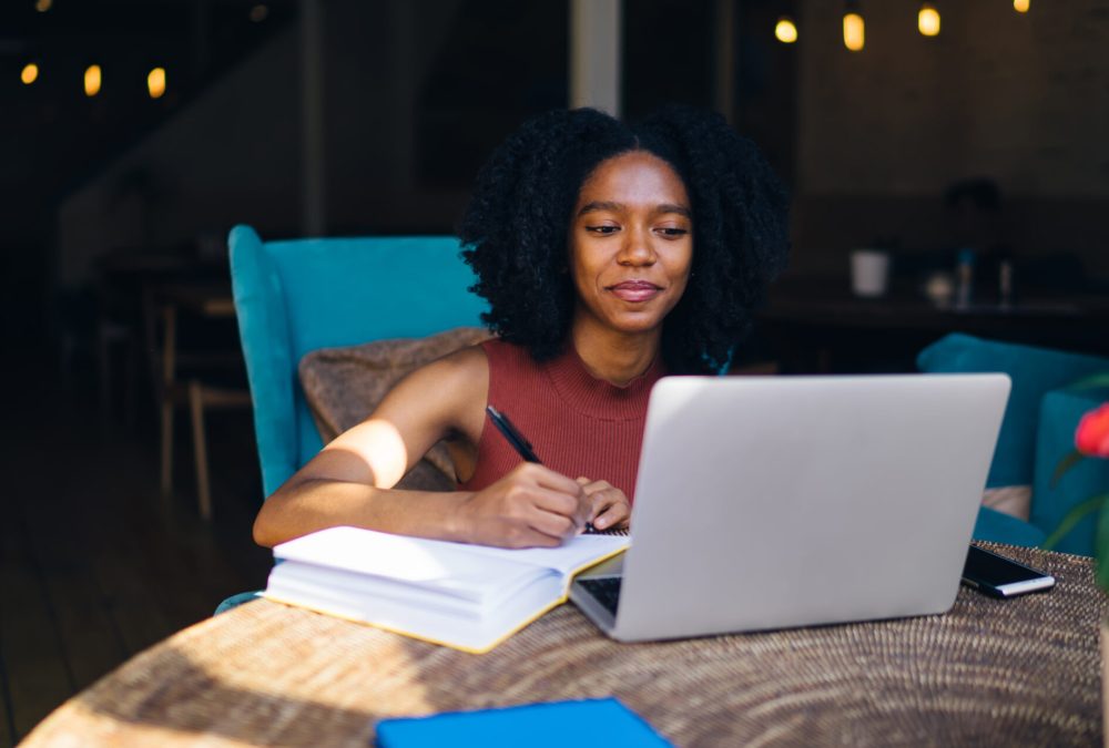 Positive dark skinned hipster girl reading questions of test for finding out features of own character on internet website and writing personal answers in textbook for education sitting at laptop