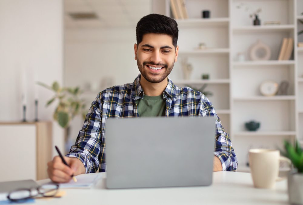 Front portrait of young smiling Arab man using laptop sitting at desk, writing in notebook. Cheerful guy browsing internet, watching webinar studying online, looking at pc screen, free copy space